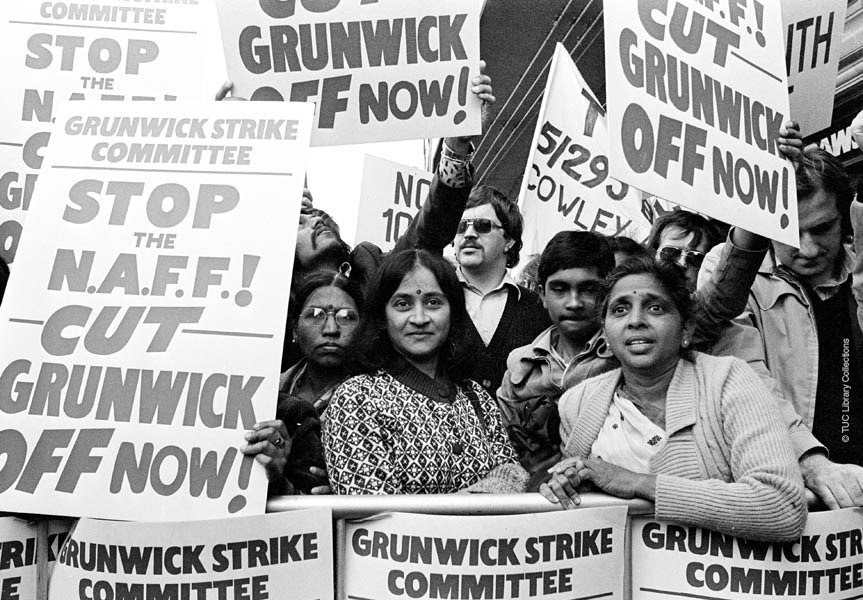 A group of women associated with the Grunwick Strike Committee protesting, some holding signs that say "GRUNWICK STRIKE COMMITTEE STOP THE N.A.F.F. CUT GRUNWICK OFF NOW!"