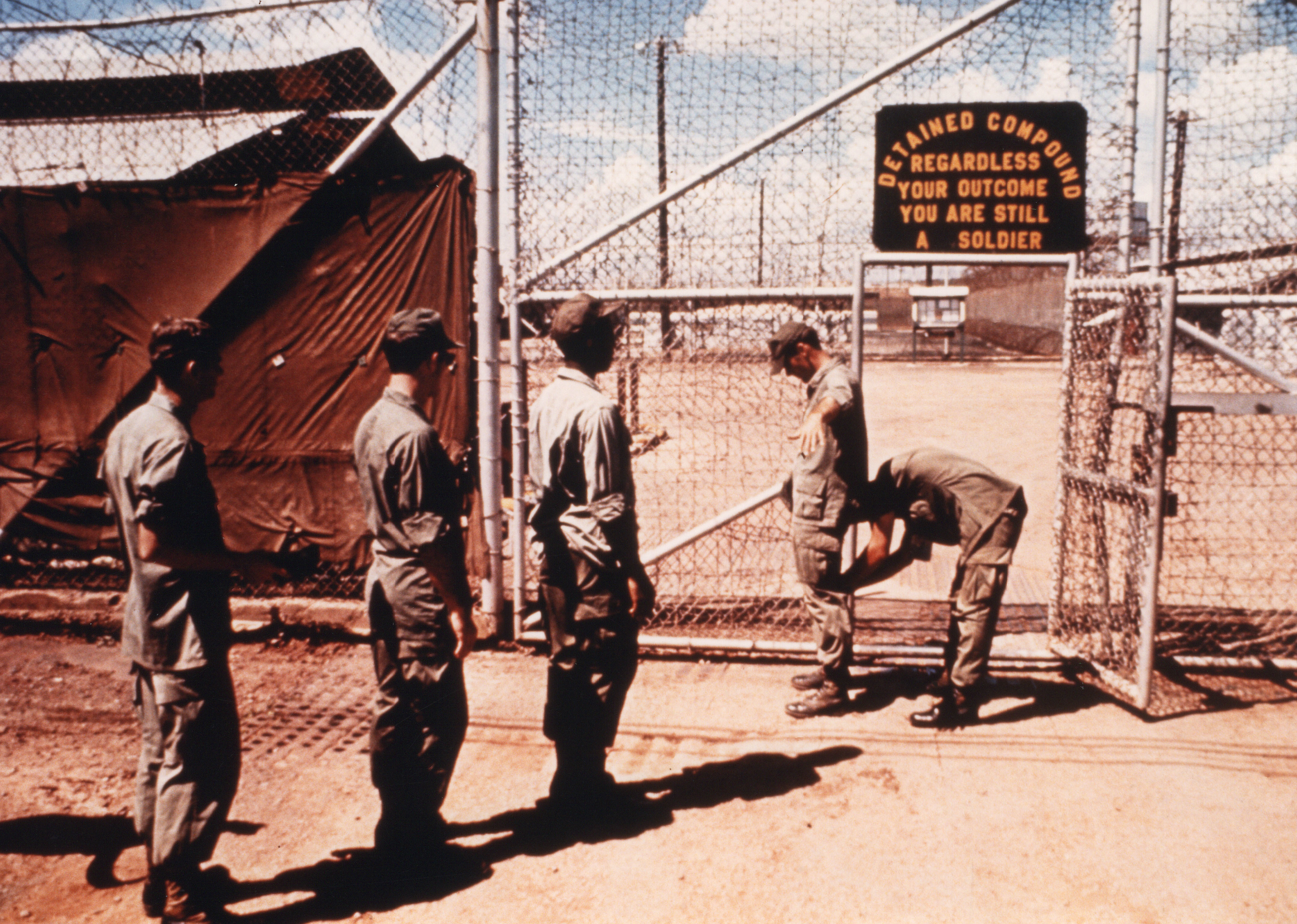 Guard searches prisoners at the gate to the pre-trial compound. A sign above the gate reads "Regardless of the outcome, you're still a soldier" [npr.org]